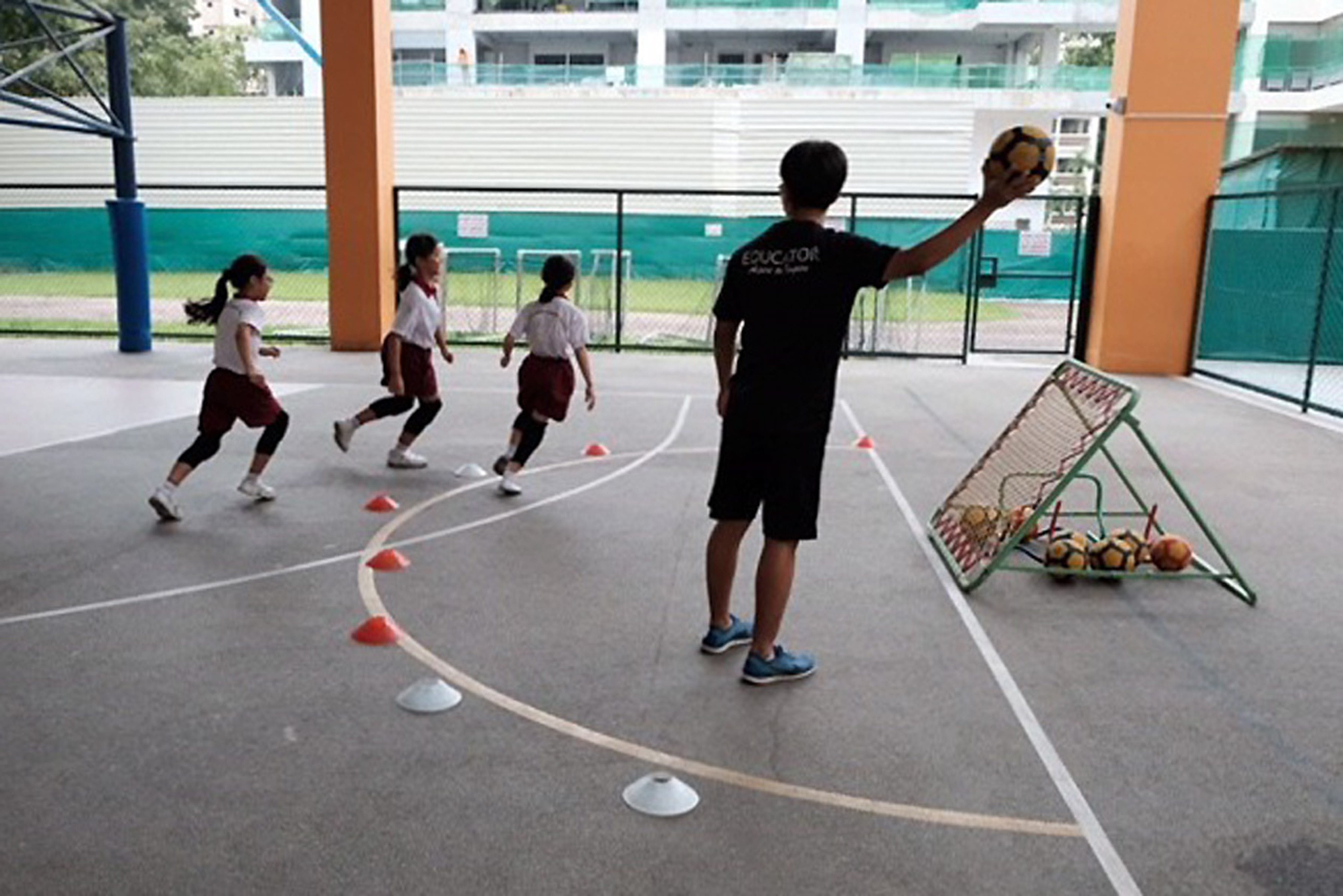 A coach holds a ball up while three children in uniforms run through cone drills on an outdoor court.
